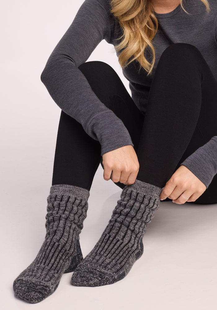 Wearing Woolx Womens Joey Crew Socks in Stone Grey, along with a long-sleeve gray shirt and black leggings, a person sits on the floor adjusting their socks. Only their torso, arms, and legs are shown—their face is not visible.