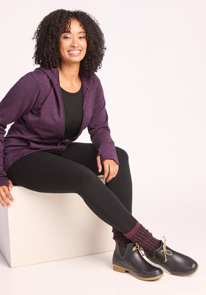 A woman with curly hair sits on a white cube, smiling. She wears a purple hoodie, black shirt and leggings, Woolx Womens Joey Crew Socks in Warm Violet, and black shoes with tan soles. The background is plain and light-colored.