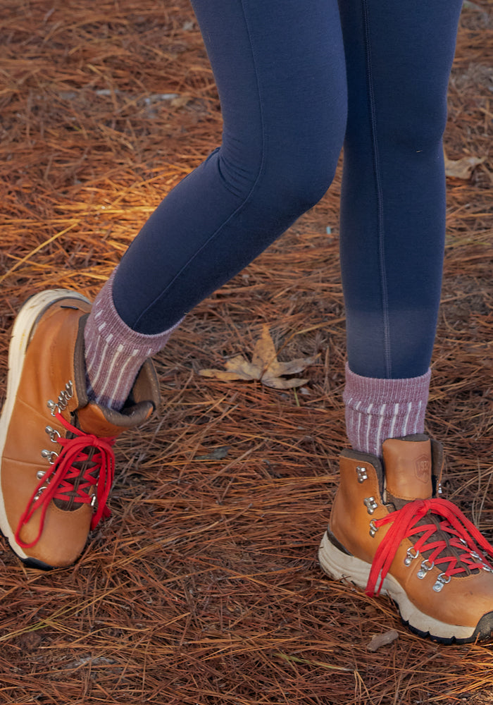 A person in navy leggings, Womens Joey Crew Socks - Wildflower by Woolx with purple stripes, and brown hiking boots with red laces stands on pine needle-covered ground.