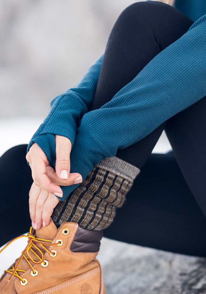 A person wearing black leggings, Woolx Womens Joey Mid Calf Socks in Dark Forest Marled, a teal long-sleeve top, and tan hiking boots sits outdoors, resting their hands on their ankle.