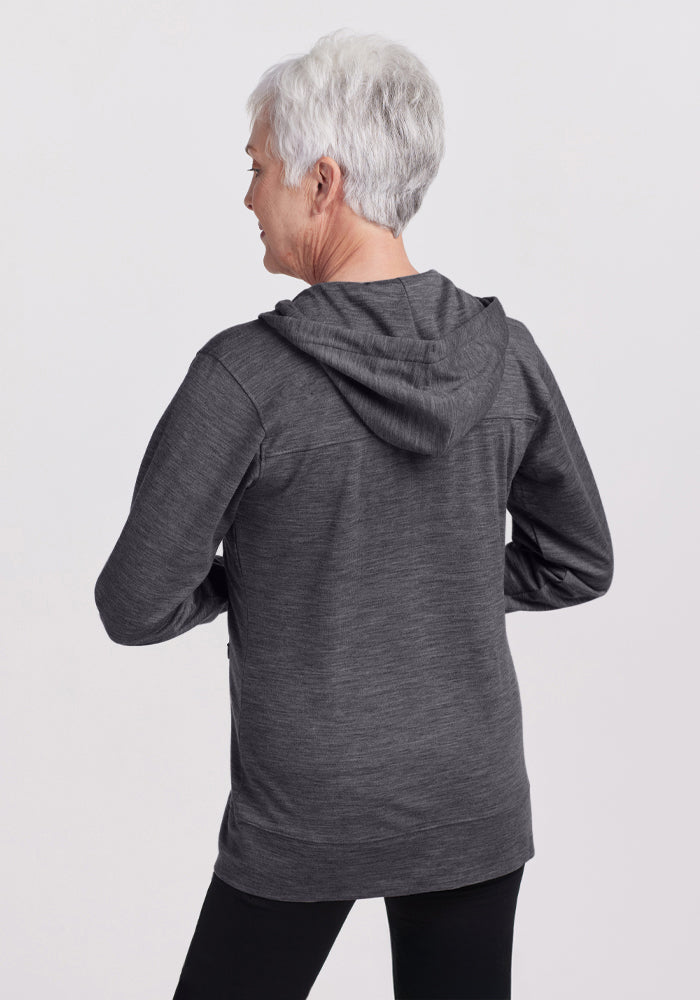 An older woman with short gray hair is shown from the back, wearing the Woolx Ryann Hoodie in Cinder Melange and black pants against a plain light background.