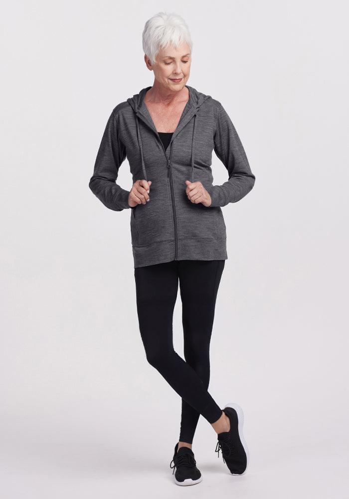 An older woman with short white hair wears the Woolx Ryann Hoodie in Cinder Melange, paired with black leggings and sneakers. She stands with one foot crossed over the other, looking down and smiling slightly against a plain light background.