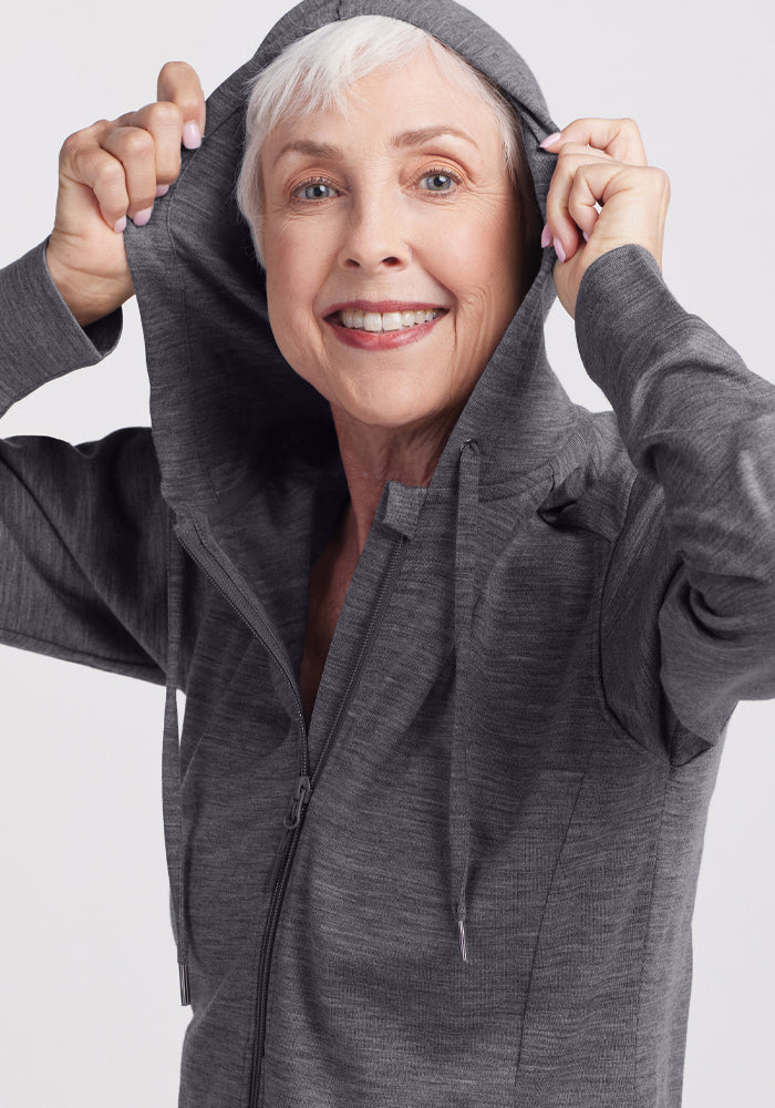 Smiling older woman with short gray hair wears the Woolx Ryann Hoodie in Cinder Melange, holding the hood with both hands against a plain light background.