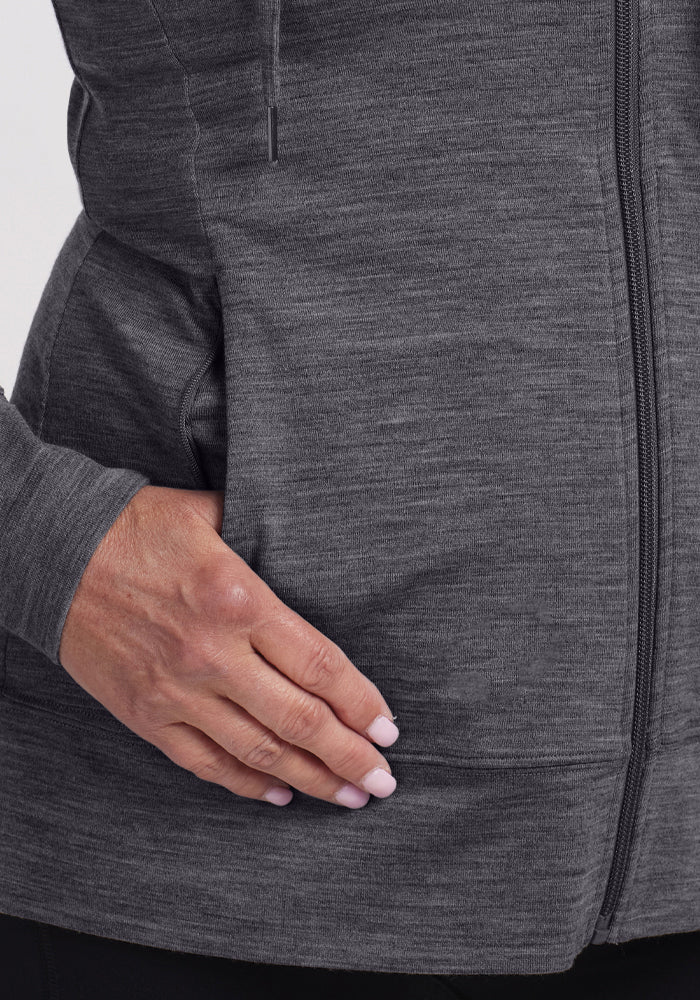 A close-up of a hand resting on the pocket of the Woolx Ryann Hoodie - Cinder Melange. The women's jacket features a heathered texture, and the person’s neatly manicured nails are painted light pink.