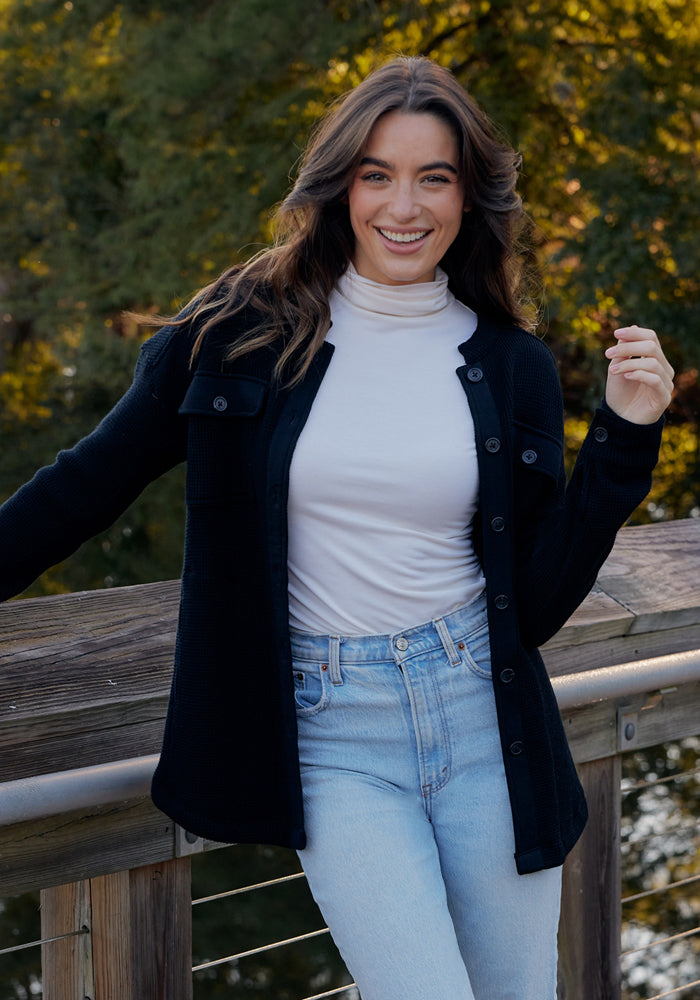 Woman with long brown hair wearing the Woolx Kennedy Bodysuit in Buttercream and a black jacket on a wooden bridge.