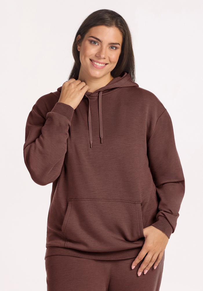 Woman smiles in a Woolx Avery Hoodie - Canyon Clay with front pocket, standing against a plain white background. 