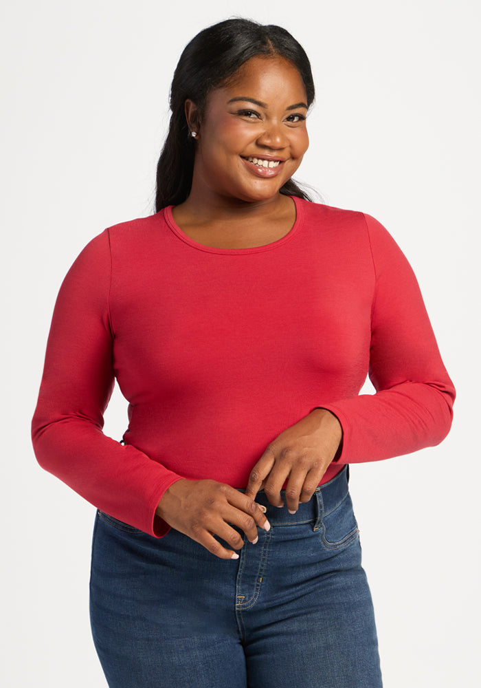 A smiling woman in a Woolx Margot Bodysuit - Maraschino Cherry and blue jeans stands against a plain white background. 