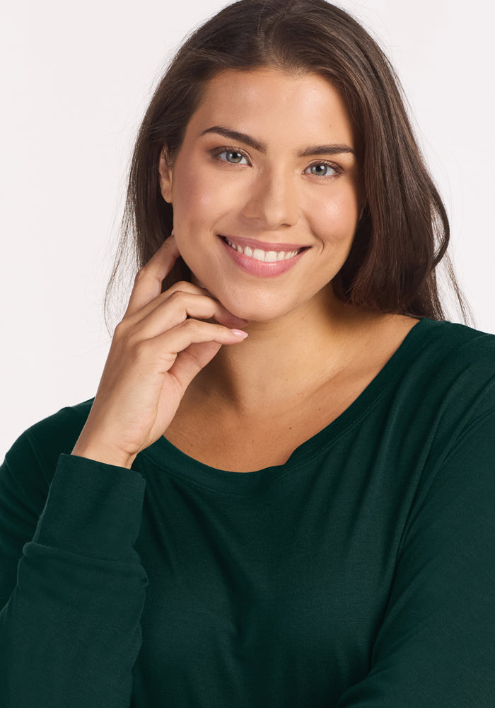 A smiling woman wearing a deep pine Woolx Ashlie Lounge Top, a long-sleeved, dark green, wrinkle-resistant merino wool top.