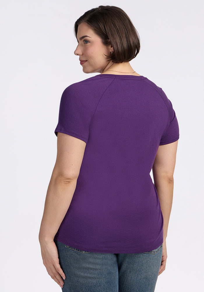 A woman with short brown hair, wearing the Woolx Mia V Neck in Plum Crush and blue jeans, stands facing away, looking over her shoulder against a plain white background.