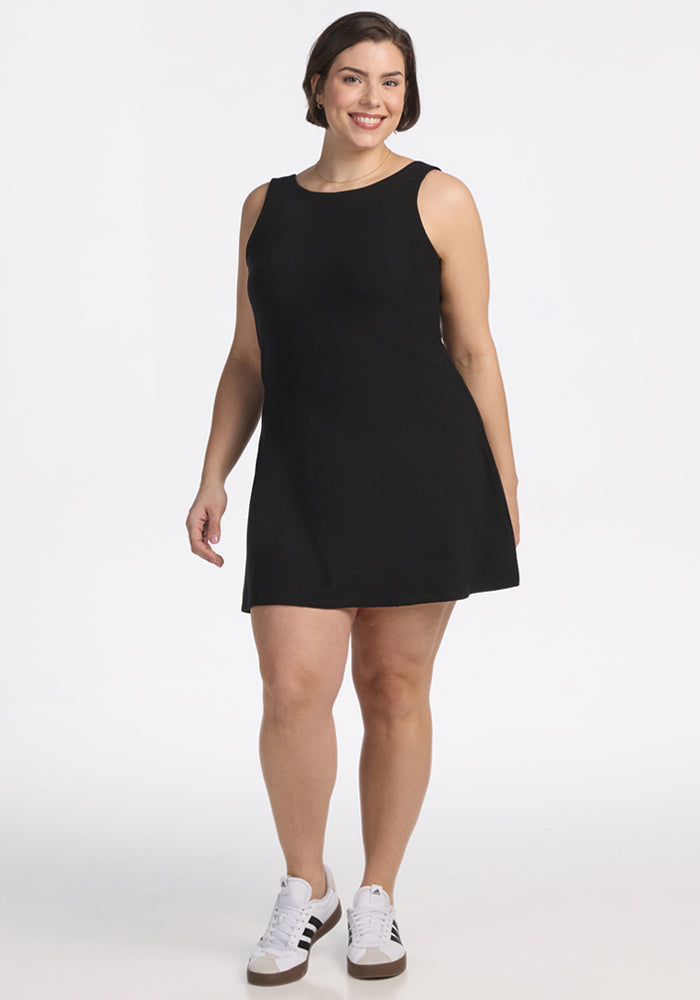 A woman with short brown hair wears the Woolx Adrienne Active Dress in black, paired with white sneakers featuring black stripes, as she stands and smiles against a plain white background.
