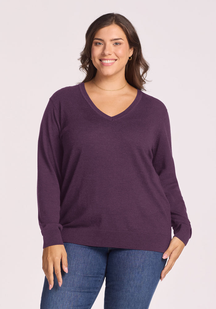 Smiling woman in a Woolx Francesca Sweater - Purple Iris and jeans stands against a plain light background. 