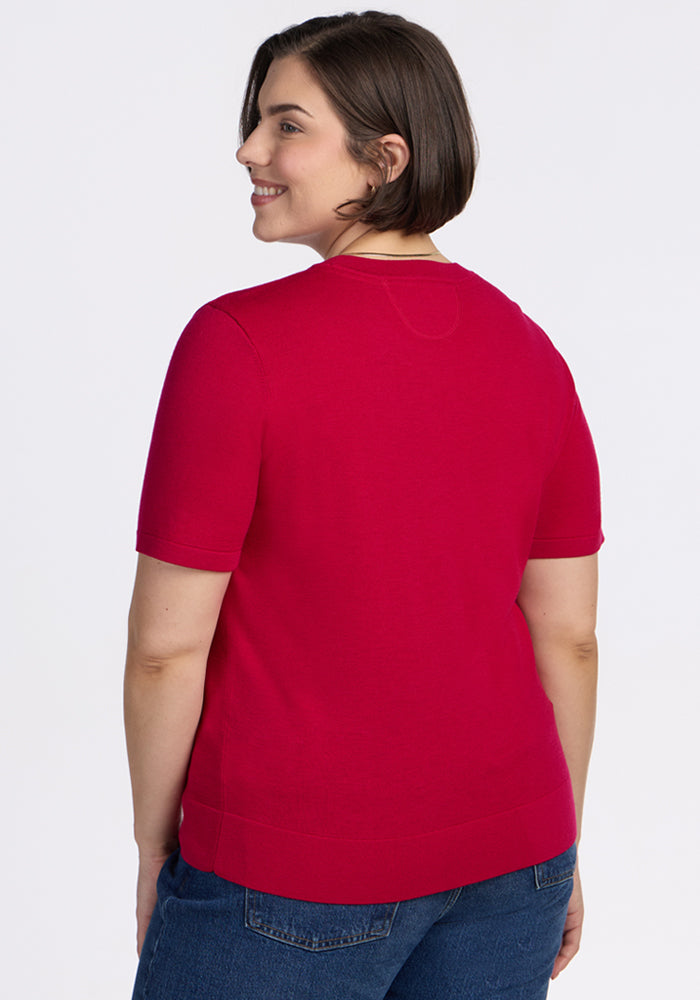 A person with short brown hair smiles over their shoulder, wearing the Woolx Anna Crew Sweater in Poppy Spark and blue jeans against a plain white background.