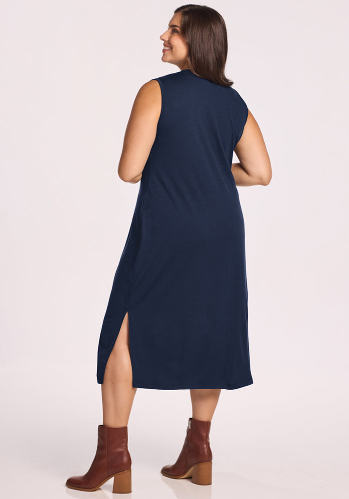 A woman with shoulder-length brown hair smiles while wearing the Woolx Cassie Dress in Deep Navy—a sleeveless, wrinkle-resistant Merino wool dress with a side slit—paired with brown heeled ankle boots against a plain light background.