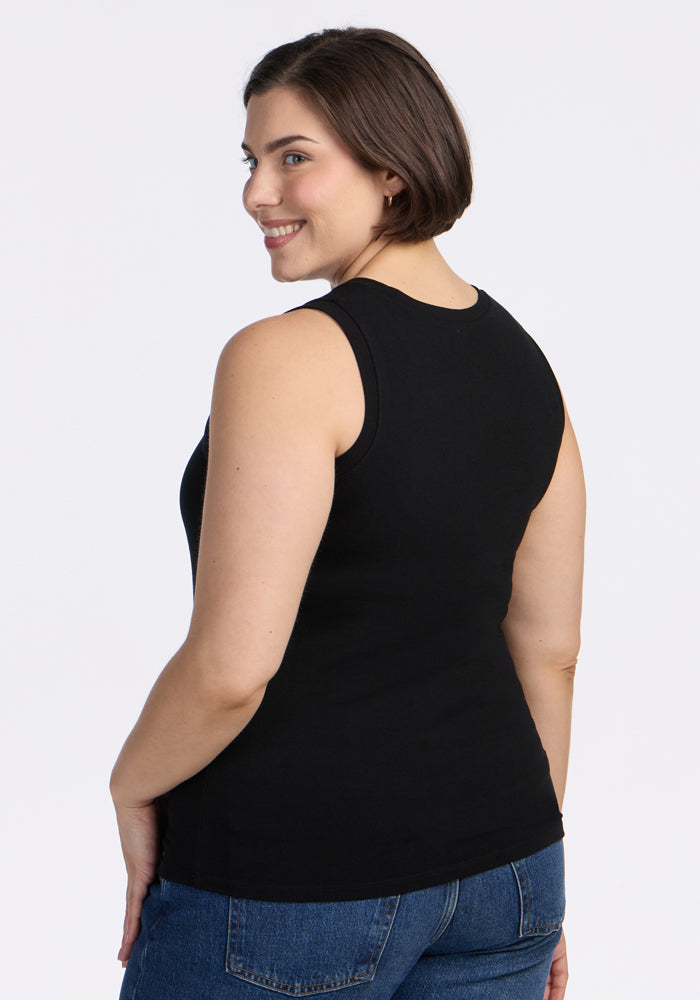 A woman with short brown hair wears the Woolx Ava Tank Top in black and blue jeans, standing slightly turned and smiling over her shoulder against a plain white background.