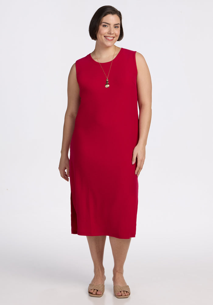A woman with short brown hair smiles while wearing the Woolx Iris Ribbed Dress in Poppy Spark, a long necklace, and beige sandals. She stands against a plain white background. 