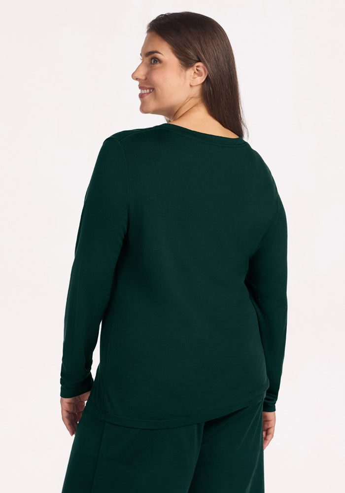 A woman seen from the back wearing a long-sleeved, deep green Lily V Neck Lounge Top - Deep Pine merino wool sleep shirt.