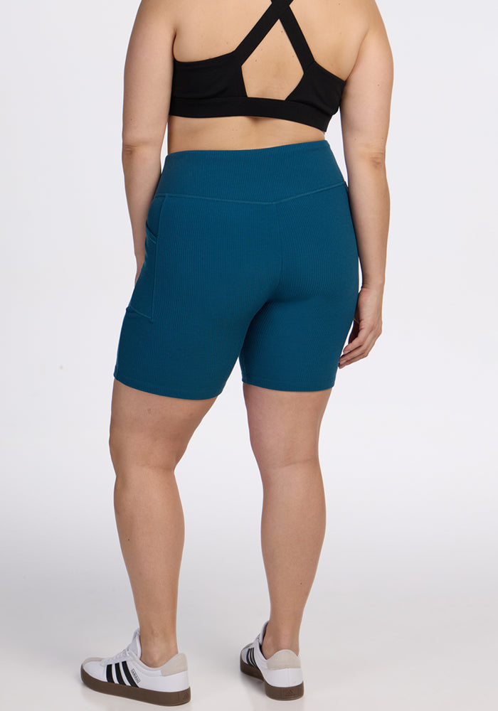 A person is shown from the back against a plain white background wearing a black sports bra, Woolx Miley Bike Shorts in Moroccan Blue, and white sneakers with black stripes.