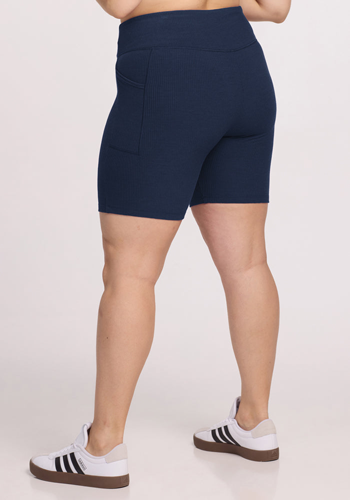 A person shown from the back wears Woolx Miley Bike Shorts in Deep Navy, featuring a high-waisted, ribbed design with pockets, paired with white Adidas sneakers with black stripes, standing against a plain light background.