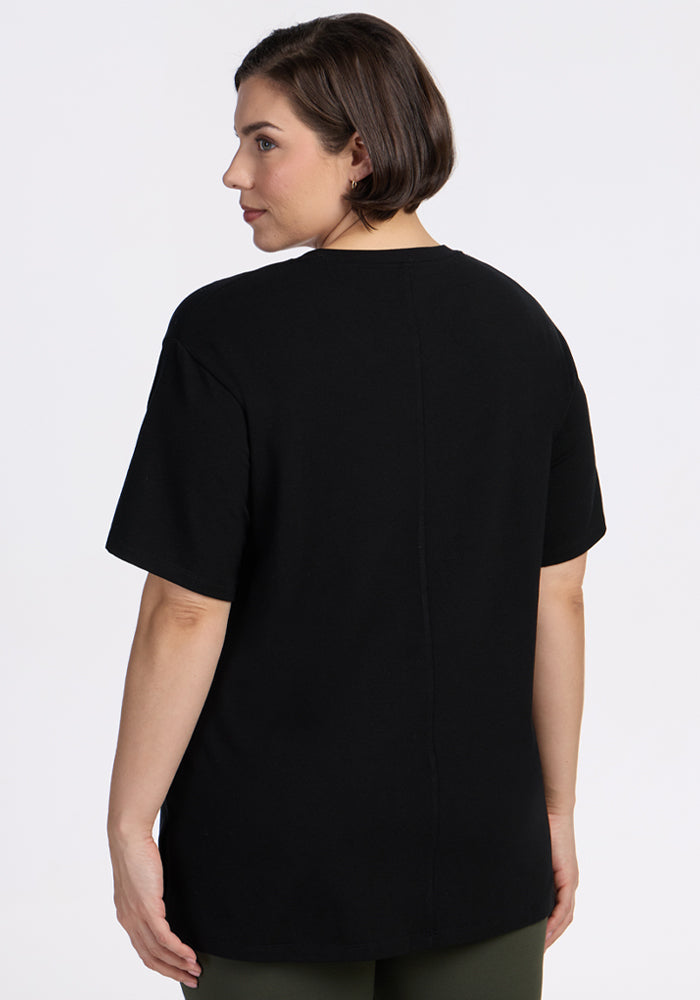 A woman with short brown hair is seen from behind, wearing the Woolx Audrey Tee in black paired with olive green pants, standing against a plain white background.