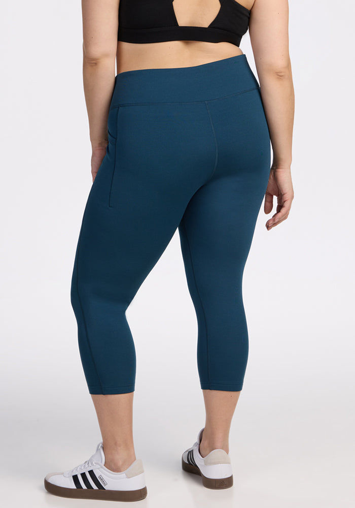 A person stands with their back to the camera, wearing Woolx Merino Flex® McKenna Capris in Majolica Blue, a black sports bra, and white-black athletic shoes against a light background.