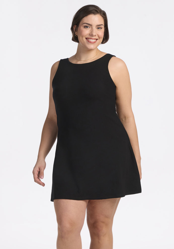 A woman with short brown hair smiles in the Woolx Adrienne Active Dress - Black, a sleeveless knee-length dress with built-in shorts, standing against a plain white background. 