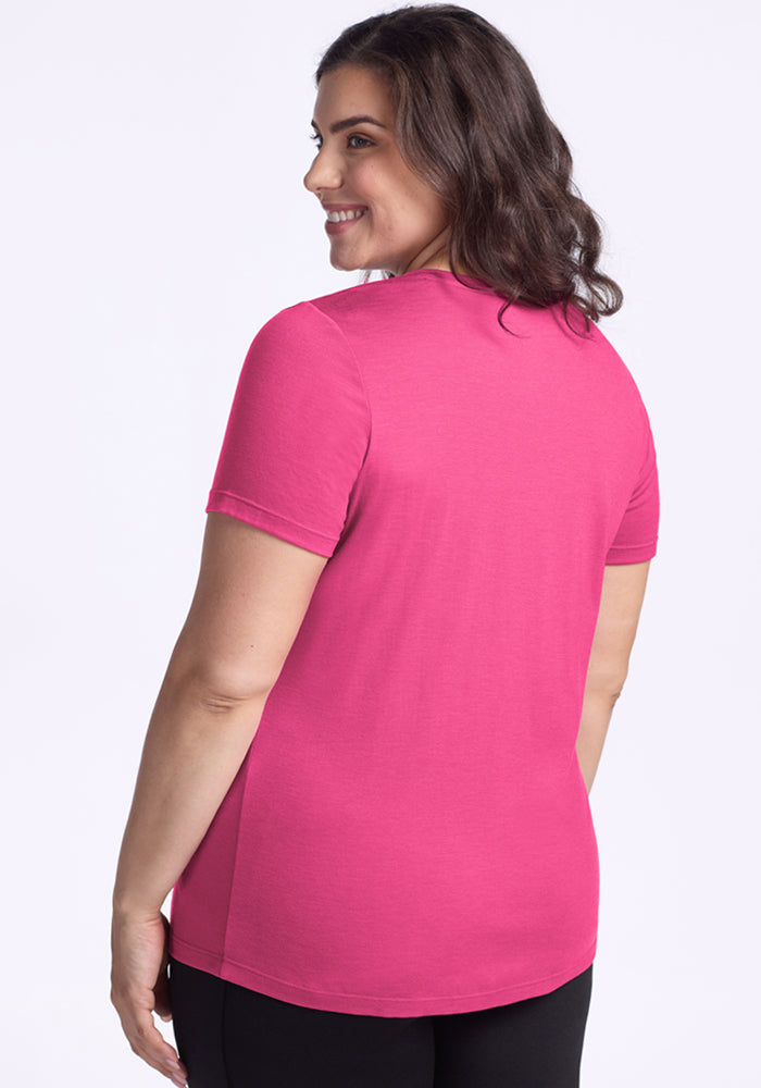 A woman with wavy brown hair stands smiling away from the camera, wearing the Woolx Junie V Neck in Raspberry Fizz and black pants, set against a plain light background.