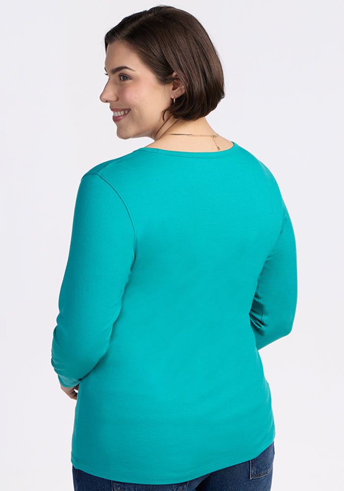 A woman with short brown hair stands smiling with her back to the camera, wearing the Woolx Remi Long Sleeve T-Shirt in Cool Jade and blue jeans against a plain, light background.