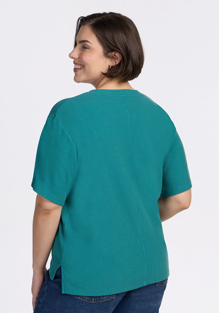 A person with short brown hair smiles while facing away from the camera, wearing the Woolx Delia Ribbed Short Sleeve - Surfside and blue jeans against a plain white background.