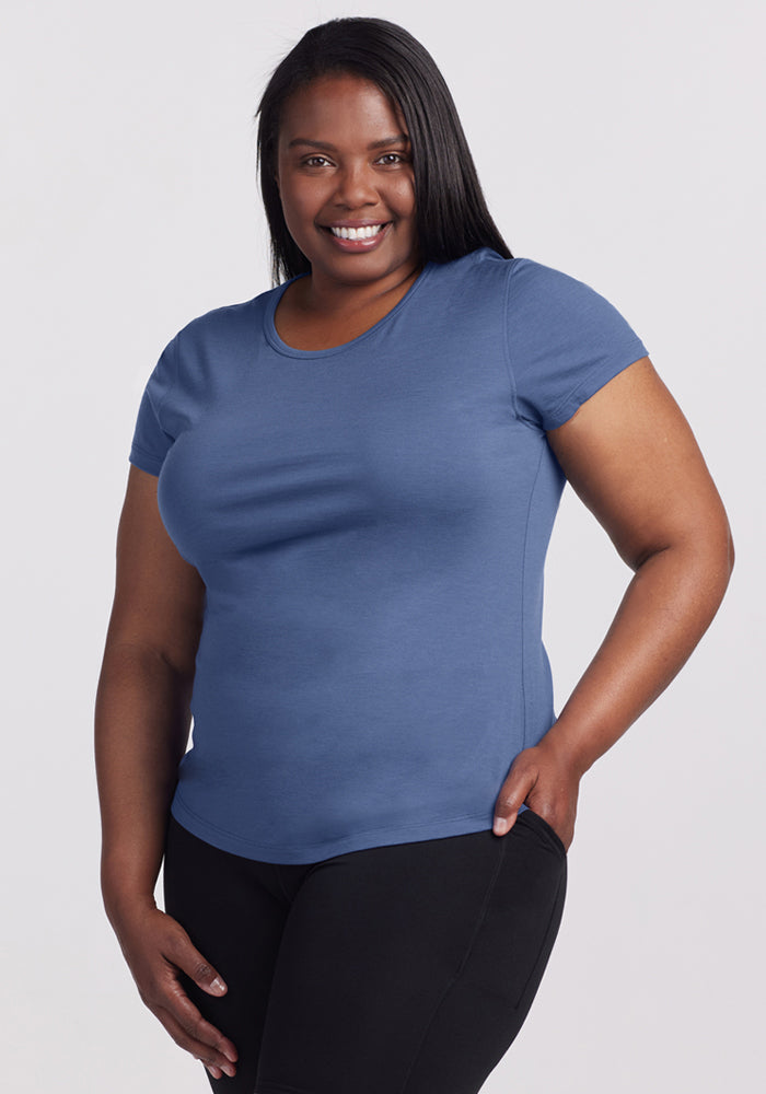 A smiling woman with long dark hair wears the Woolx Addie Crew Neck - Skyline and black pants, standing against a plain light background with one hand on her hip. 