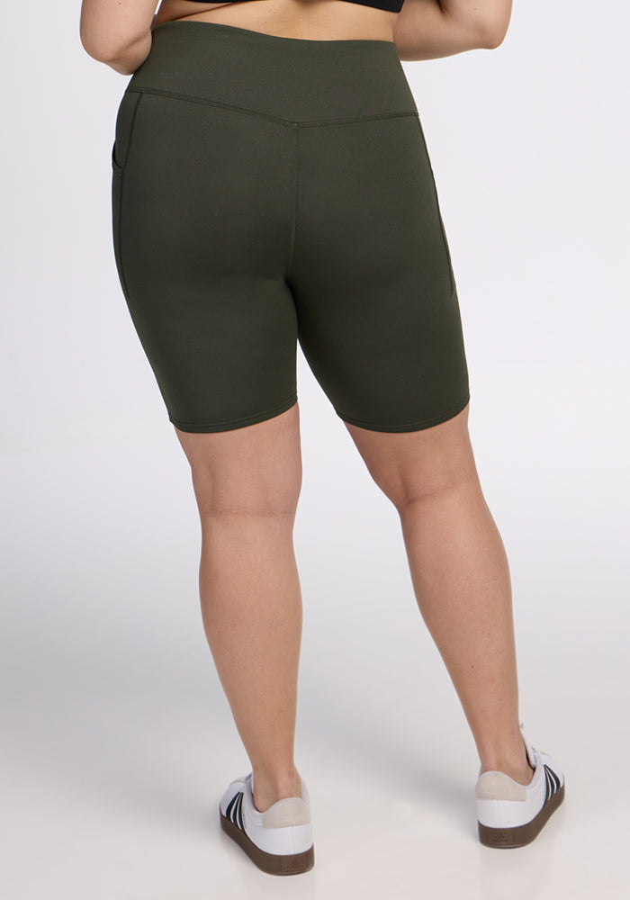 A person stands with their back to the camera, wearing Woolx Merino Flex® Dani Bike Shorts in Everleaf and a black top, paired with white sneakers featuring black stripes against a plain white background.