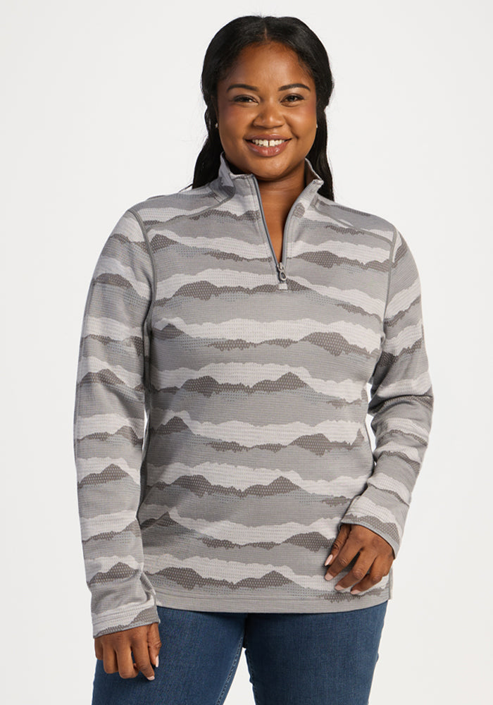 Smiling woman in Woolx Emi 1/4 Zip - High Peaks thermal top and jeans, standing against a plain background. 