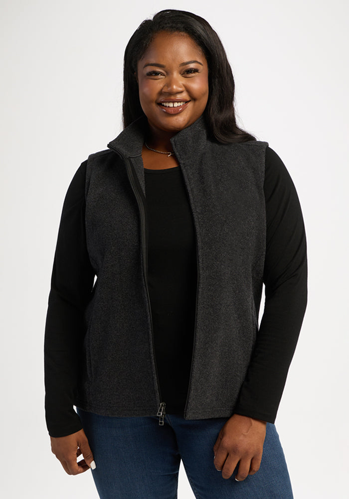 Smiling woman wears Woolx Haley Vest - Carbon Black over a black long-sleeve shirt and blue jeans, white background. 
