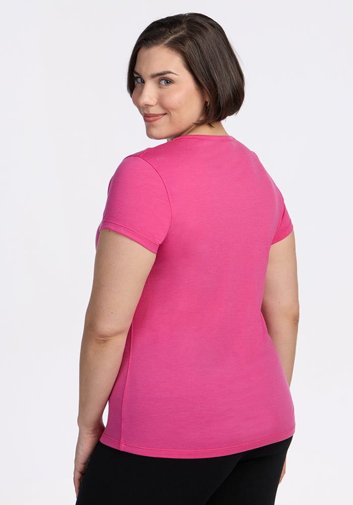 A person with short brown hair, dressed in Woolx’s Addie Short Sleeve Crew in Raspberry Fizz and black pants, stands slightly turned to the side and smiles at the camera against a plain white background.