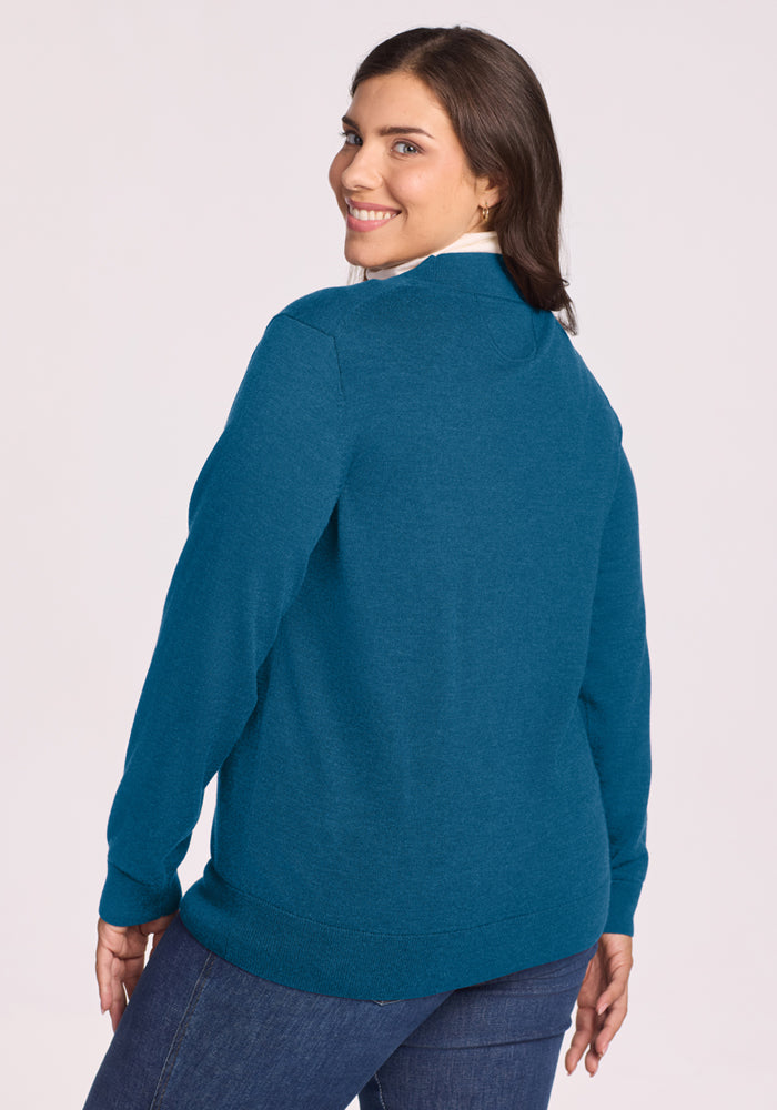 Woman in Woolx Gia Sweater - Blue Pine and jeans smiles over her shoulder against a plain light background.