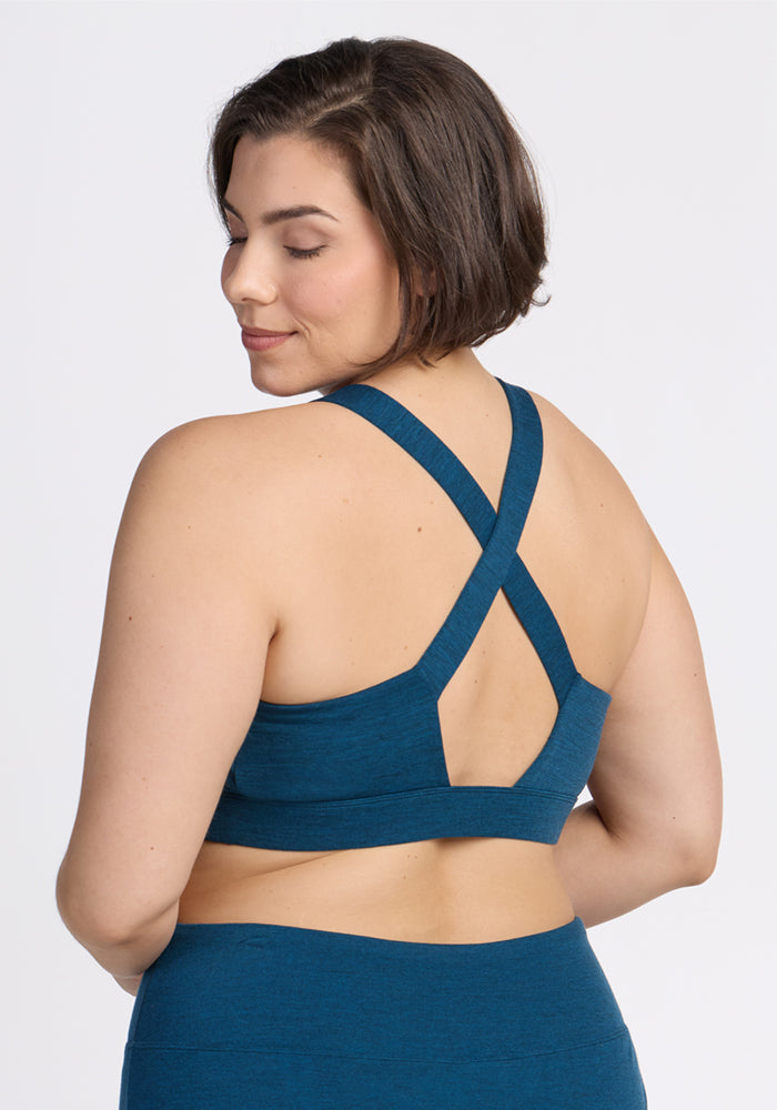 A woman with short brown hair is shown from the back, wearing the Woolx Livi Bra in Majolica Blue with crisscross straps and matching leggings. She stands against a white background, her slight smile hinting at all-day comfort.