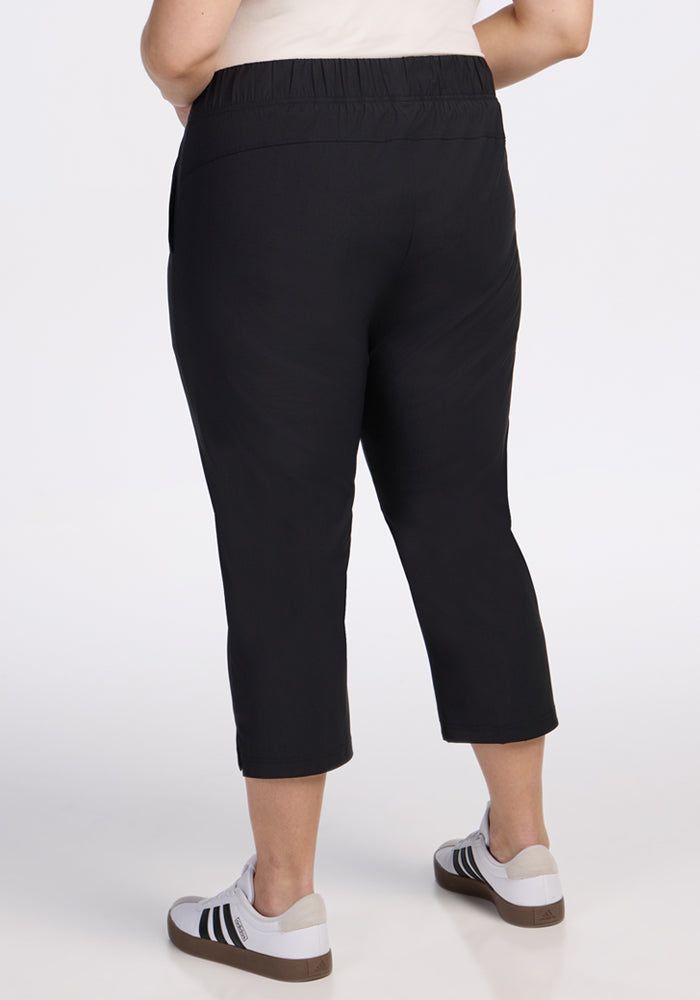 A person wearing Woolx Nicole Crop Pants in black and white sneakers stands facing away from the camera, showing the back view of the pants against a plain white background.