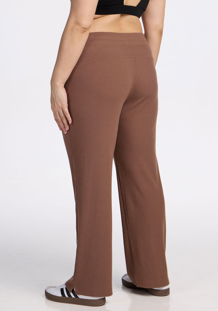 A person wearing a black crop top, Woolx Luca Ribbed Pants in Sienna Kiss, and black-and-white slide sandals stands with their back to the camera against a plain white background.