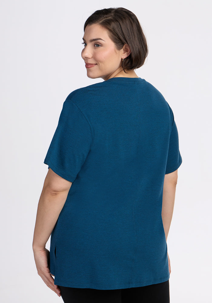A woman with short brown hair stands facing away, looking over her shoulder, wearing the Woolx Audrey Tee in Majolica Blue and black pants, set against a plain light background.