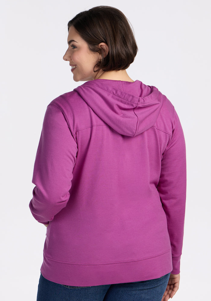 A woman with short brown hair smiles while facing away from the camera, wearing the Woolx Ryann Hoodie in Purple Wine and blue jeans against a plain white background—a perfect travel essential.