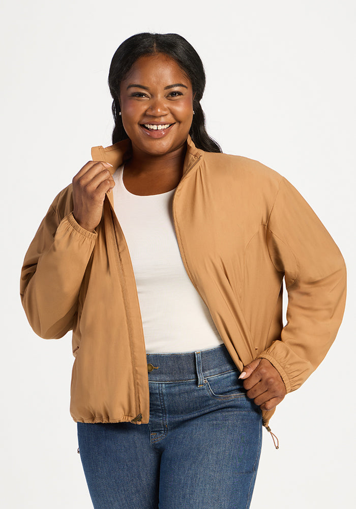 A smiling woman wears a tan Woolx Cooper Windbreaker - Saddleback over a white top and blue jeans. 
