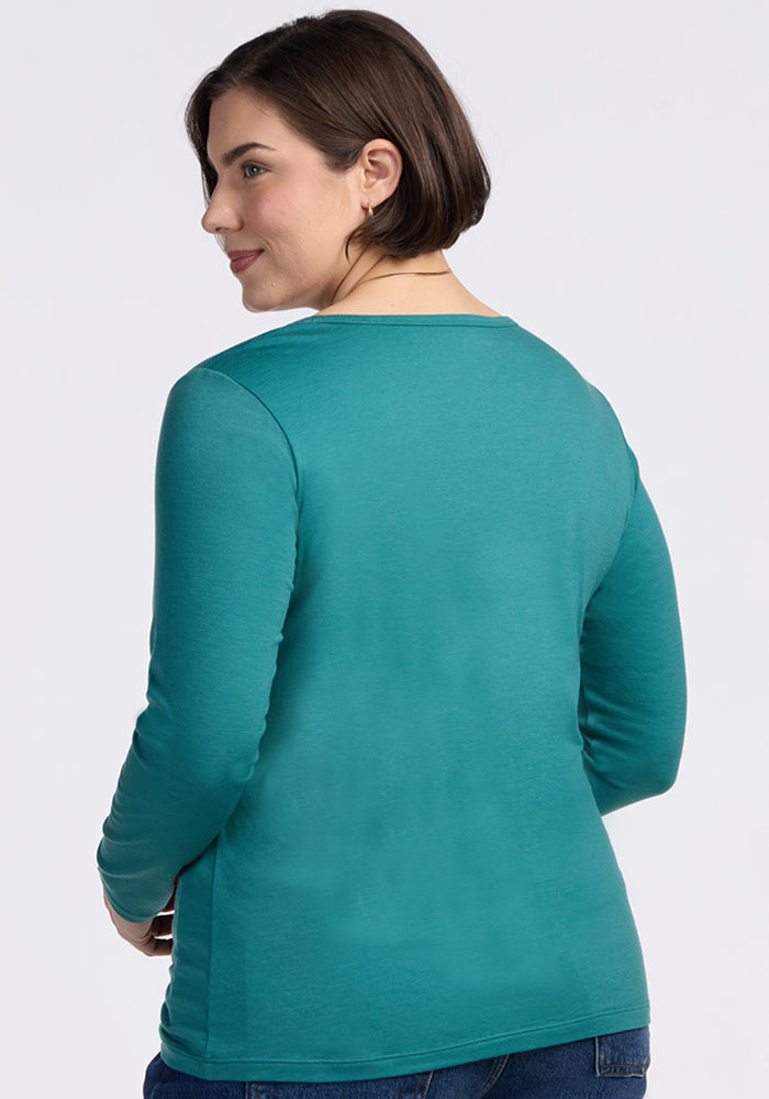 A woman with short brown hair wears the Woolx Layla V Neck Top in Surfside and blue jeans, standing facing away and looking over her shoulder with a gentle smile. The background is plain and light-colored.