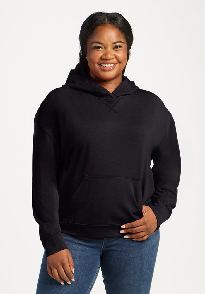 Woman smiles in a Woolx Lizzy Hoodie - Black and jeans, standing against a plain white background. 