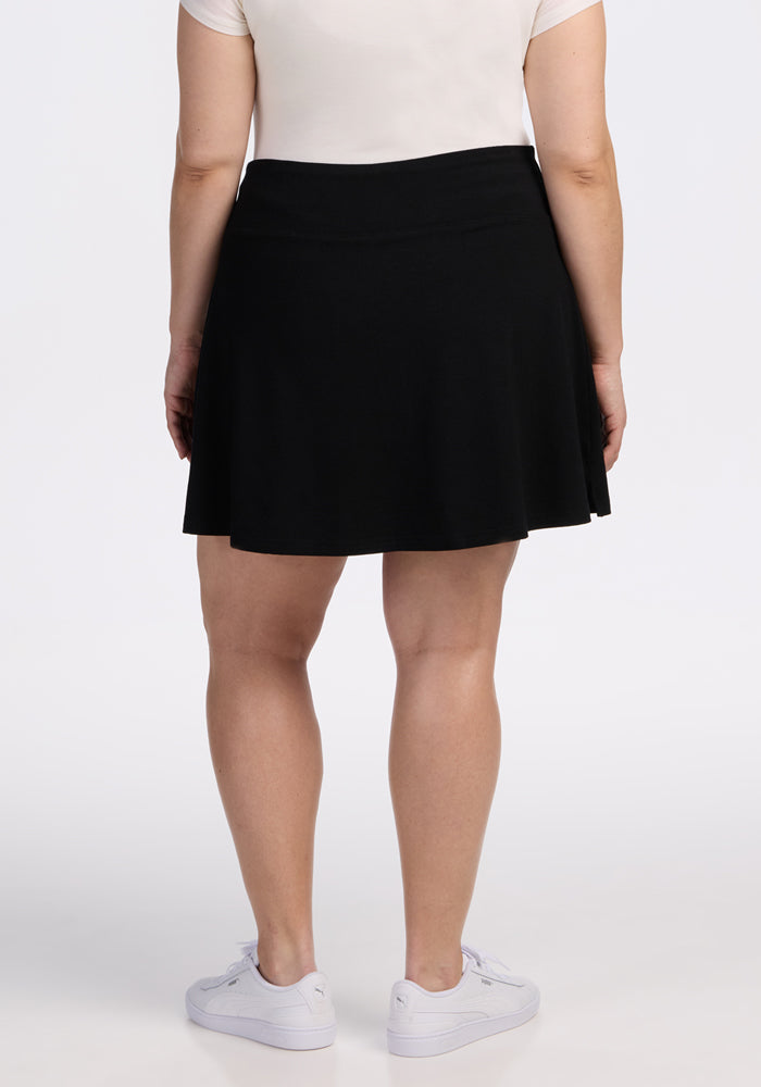 A person stands facing away from the camera, wearing a white short-sleeve shirt, Woolx Sienna Skort - Black with pockets, and white sneakers against a plain light background.