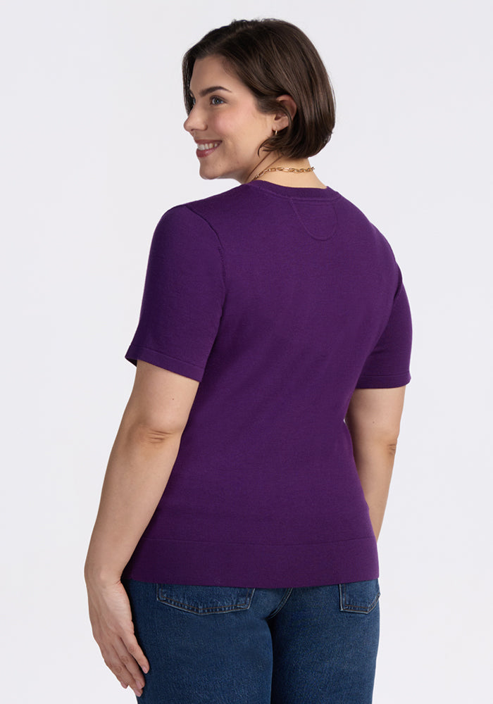 A woman with short brown hair smiles over her shoulder, wearing the Woolx Anna Crew Sweater in Plum Crush and blue jeans, standing against a plain white background.
