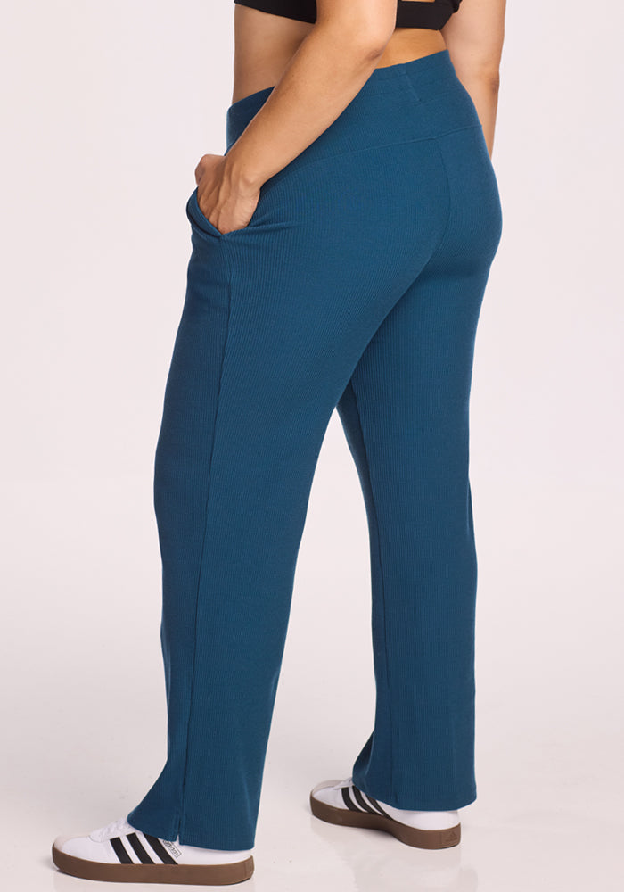 Someone wearing Woolx’s Luca Ribbed Pants in Moroccan Blue and white sneakers with black stripes stands sideways, one hand in their pocket, against a plain light background.