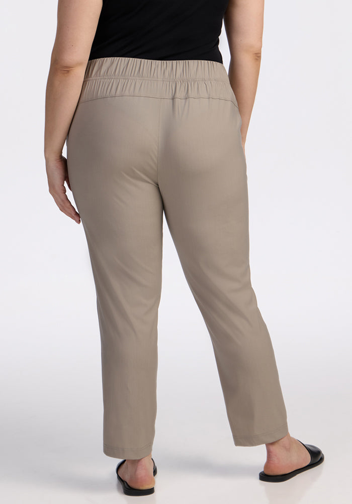 A person stands with their back to the camera wearing Woolx Nicole Ankle Pants in Vintage Khaki, a black top, and black slip-on shoes.