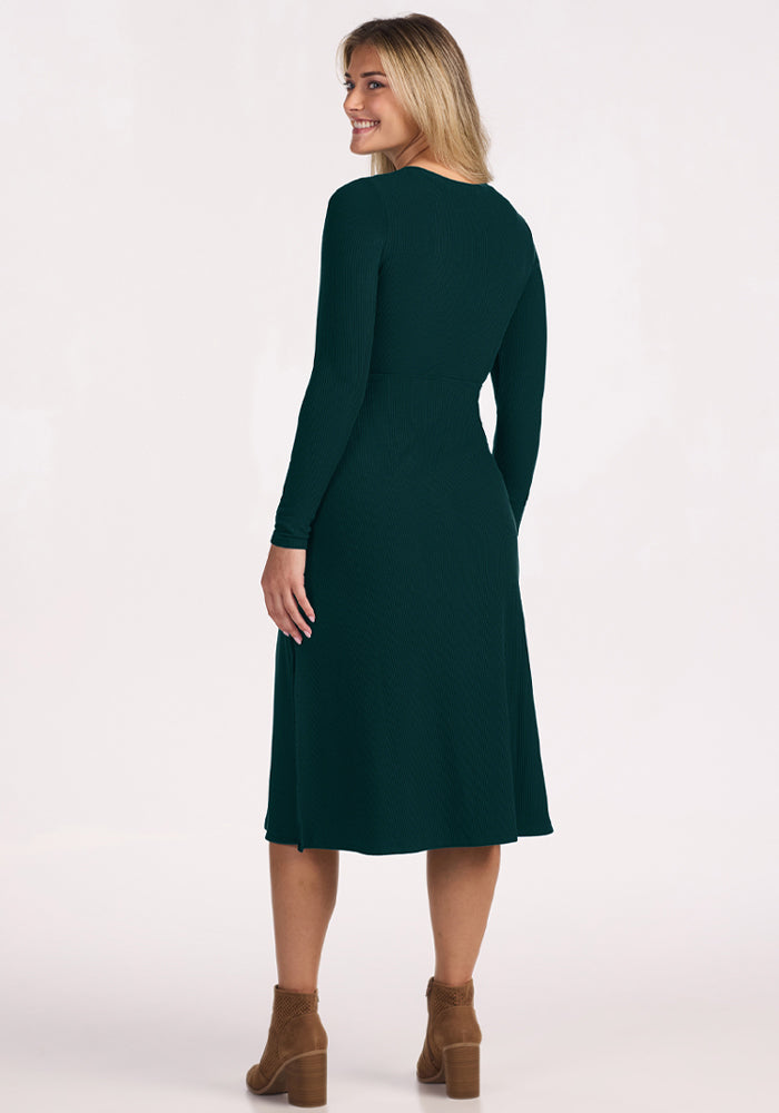 Smiling woman in Woolx Caroline Ribbed Twirl Dress—Deep Pine—stands against a plain background in brown ankle boots.