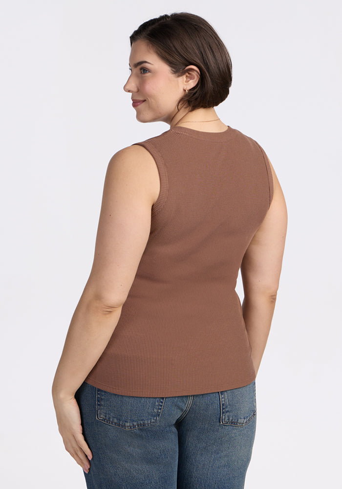 A woman with short brown hair stands against a plain white background, looking over her shoulder. She is wearing the Woolx Sloane Ribbed Tank in Sienna Kiss and blue jeans.