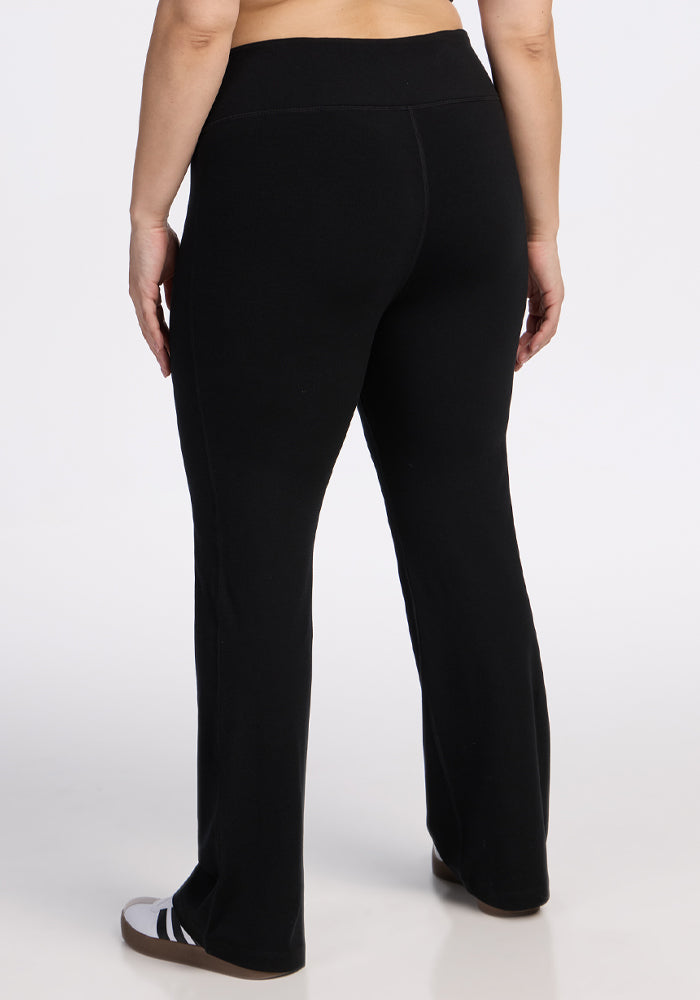 A person stands with their back to the camera, wearing Woolx Morgan Flare Pants in black and black-and-white sandals against a plain white background.