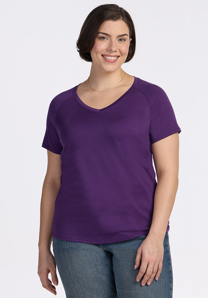 A woman with short dark hair smiles while wearing the Woolx Mia V Neck in Plum Crush and blue jeans, standing against a plain white background. 